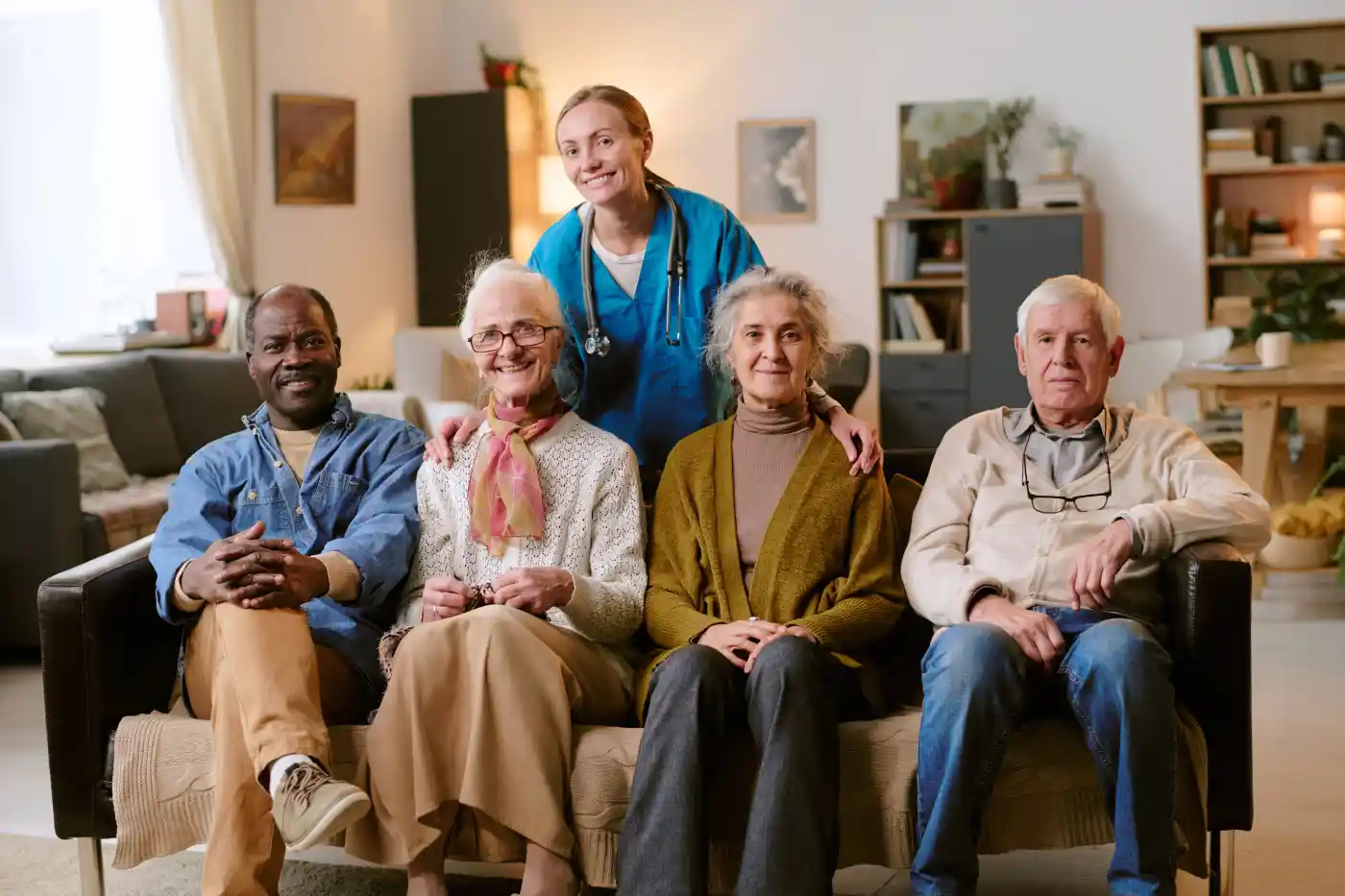 Four seniors sit on a couch with a healthcare worker standing behind them in a well-lit living room, all facing the camera—showcasing the warmth and trust that answer the question, "Why Us?.