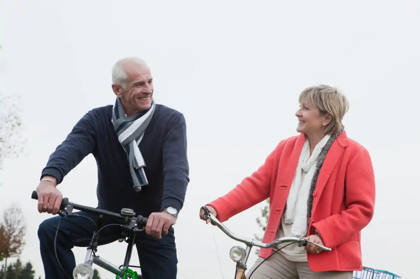 An older man and woman, both smiling, ride bicycles outdoors on a cloudy day while looking at each other—enjoying the active lifestyle supported by Upper Arlington Home Care.