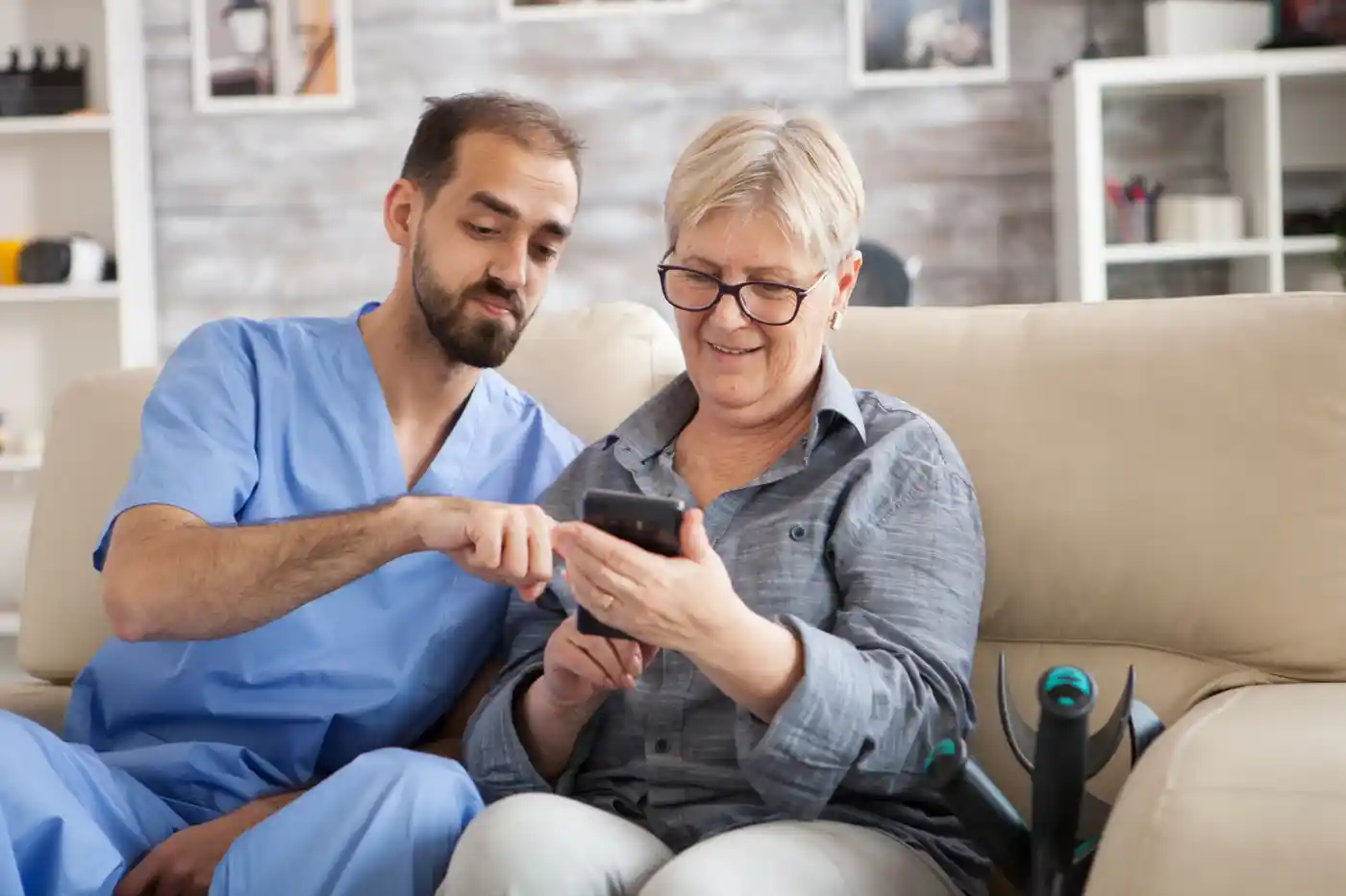 A healthcare worker in scrubs sits on a couch next to an older woman, showing her something on a smartphone.