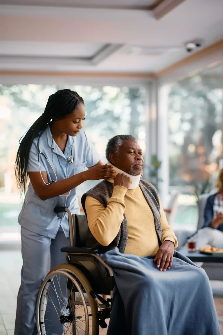 A healthcare worker adjusts a neck brace on an older adult sitting in a wheelchair, covered with a blanket, in a well-lit room where medication management services are also provided.