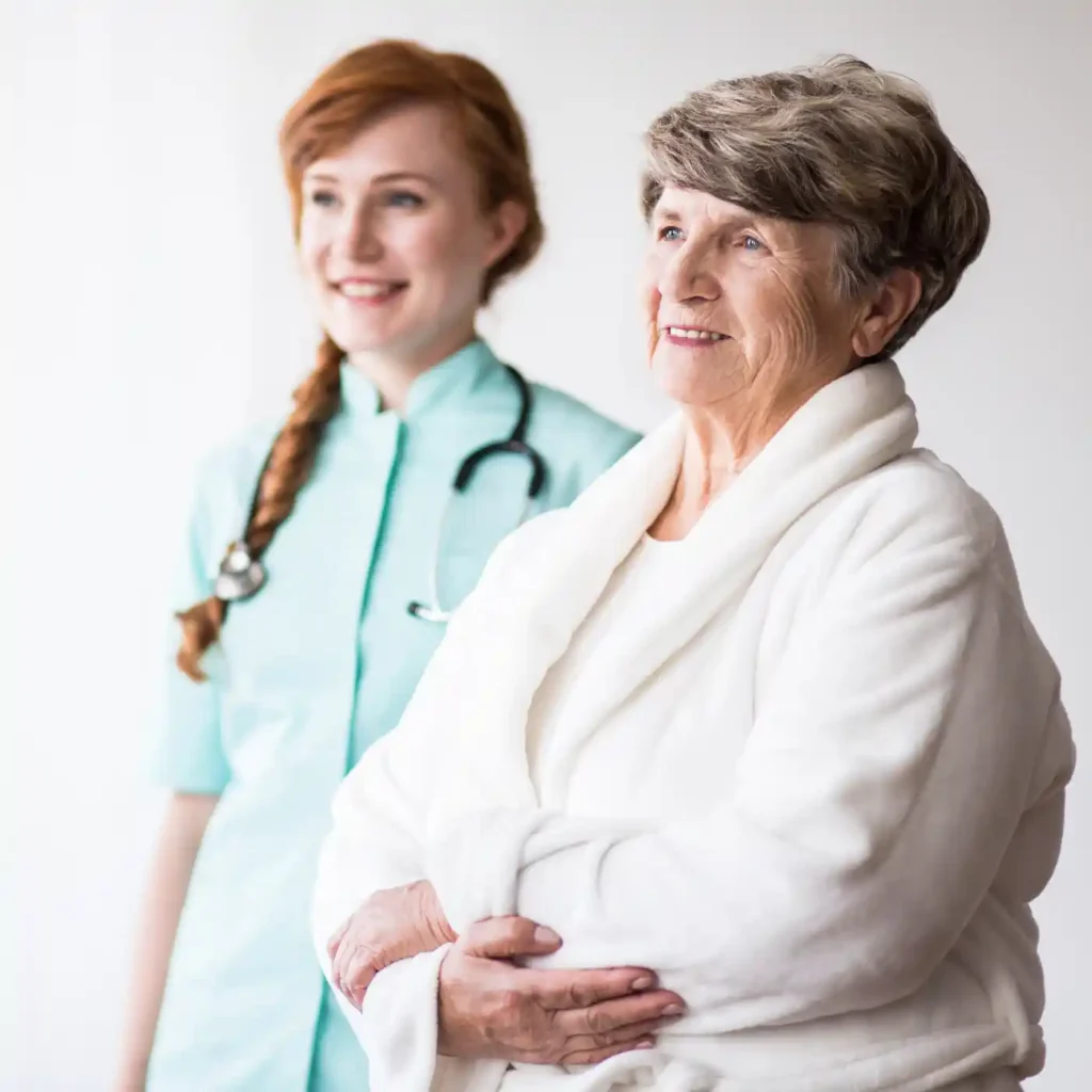 An older woman in a white robe stands beside a smiling Upper Arlington Home Care professional in a light blue uniform with a stethoscope.