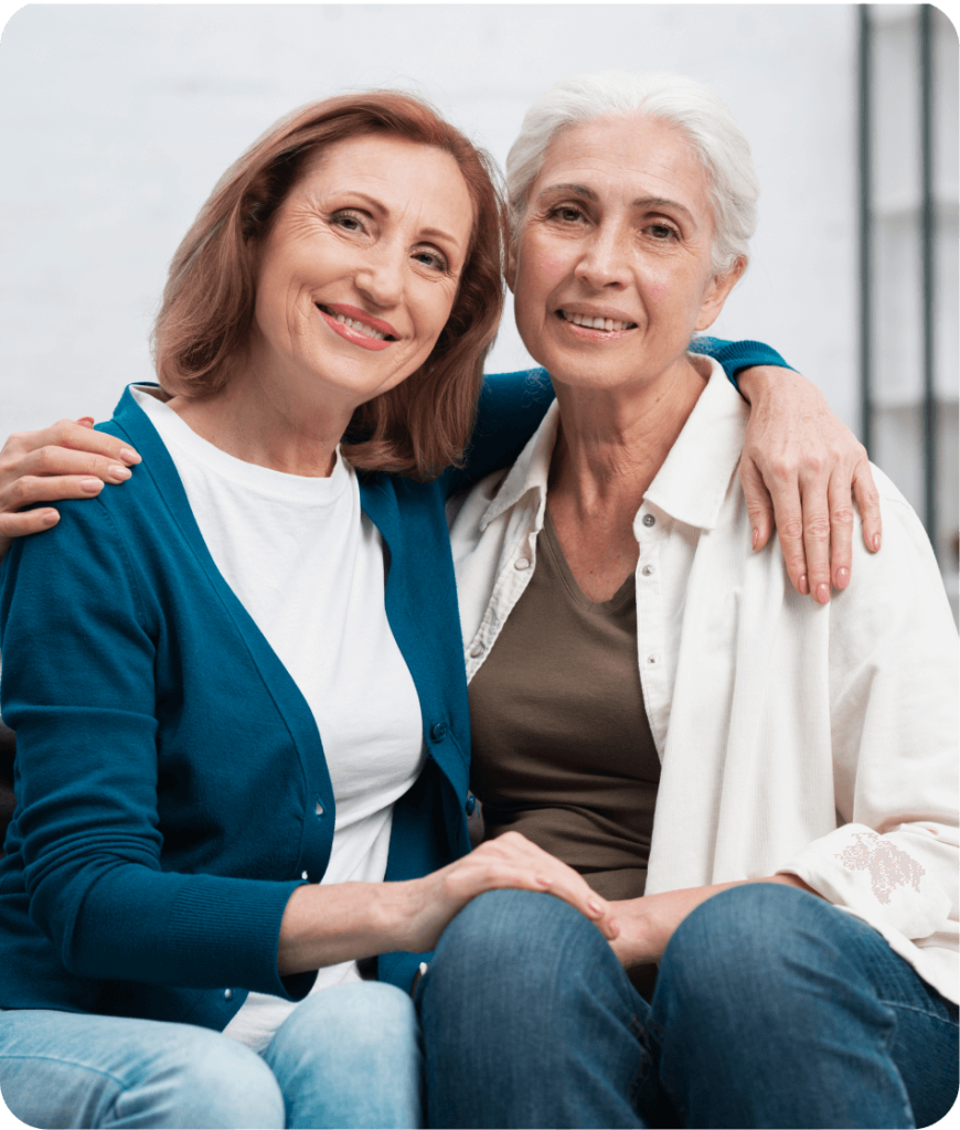 Two older women sitting close together, smiling, with one woman's arm around the other's shoulders in a casual indoor setting.