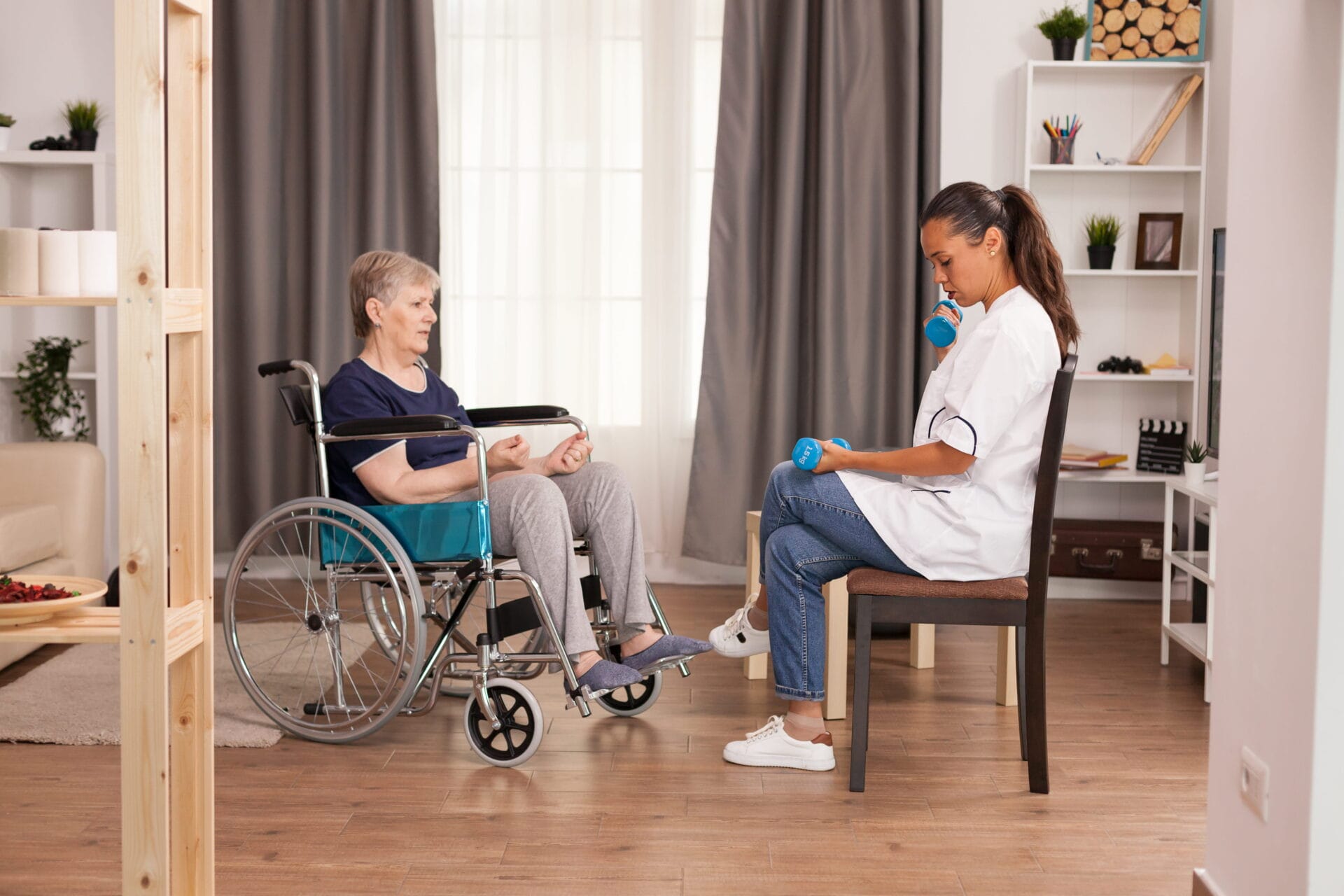 A healthcare worker sits facing an older woman in a wheelchair, demonstrating an exercise with blue dumbbells in a well-lit living room.
