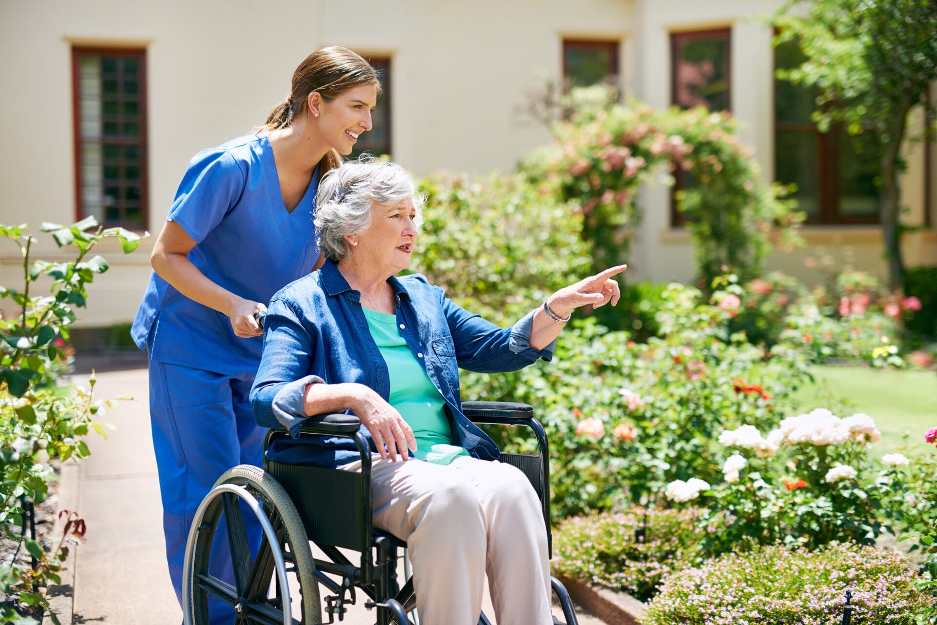 A caregiver in blue scrubs pushes an older woman in a wheelchair through a garden; the woman is pointing at something while they both smile.