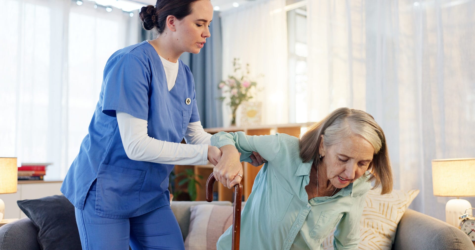 A caregiver in blue scrubs assists an elderly woman with a cane as she stands up from a couch in a well-lit living room.