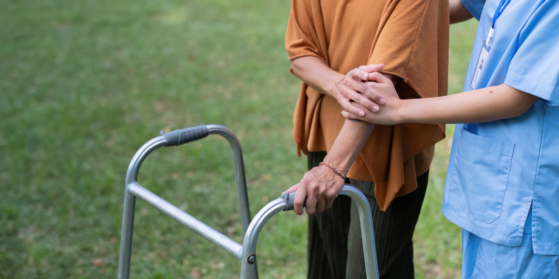 A caregiver supports an elderly person using a walker outdoors on grass.