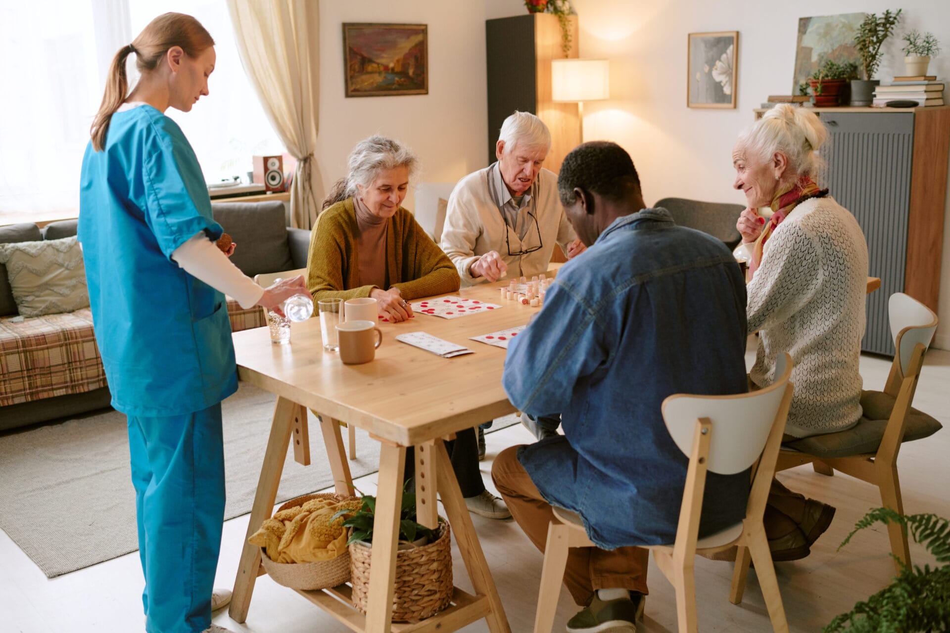 Four older adults sit around a table playing bingo while a caregiver in blue scrubs stands nearby in a cozy living room setting.