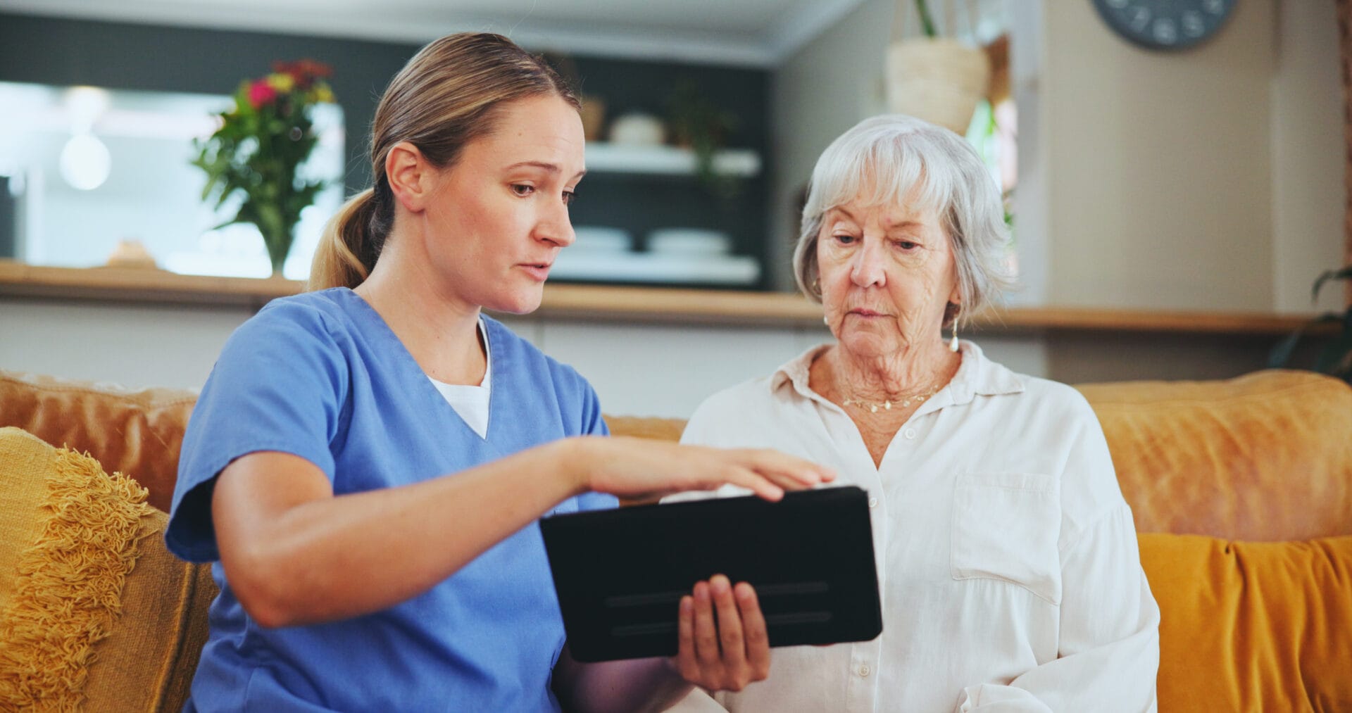 A nurse in scrubs sits on a couch showing a tablet device to an elderly woman in a white shirt in a home setting.