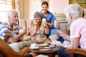 A group of older adults and a caregiver sit around a table, laughing and playing cards in a bright, cozy room.