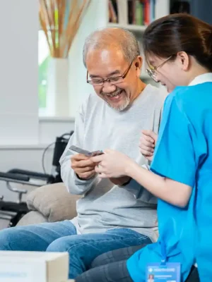 An elderly man sits on a couch smiling and looking at a card, while a healthcare worker in blue uniform—providing home care services—sits beside him. A wheelchair is visible in the background.