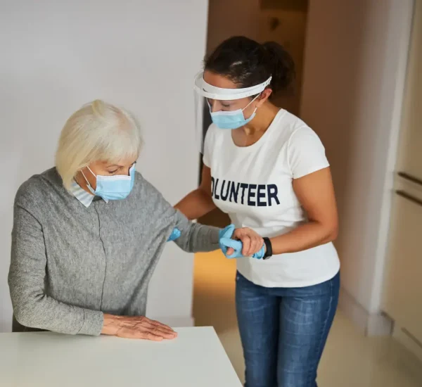 A female volunteer wearing a face shield and mask helps an elderly woman in a mask sit at a table, reflecting the compassionate spirit of Home Care Columbus in a warm home setting.