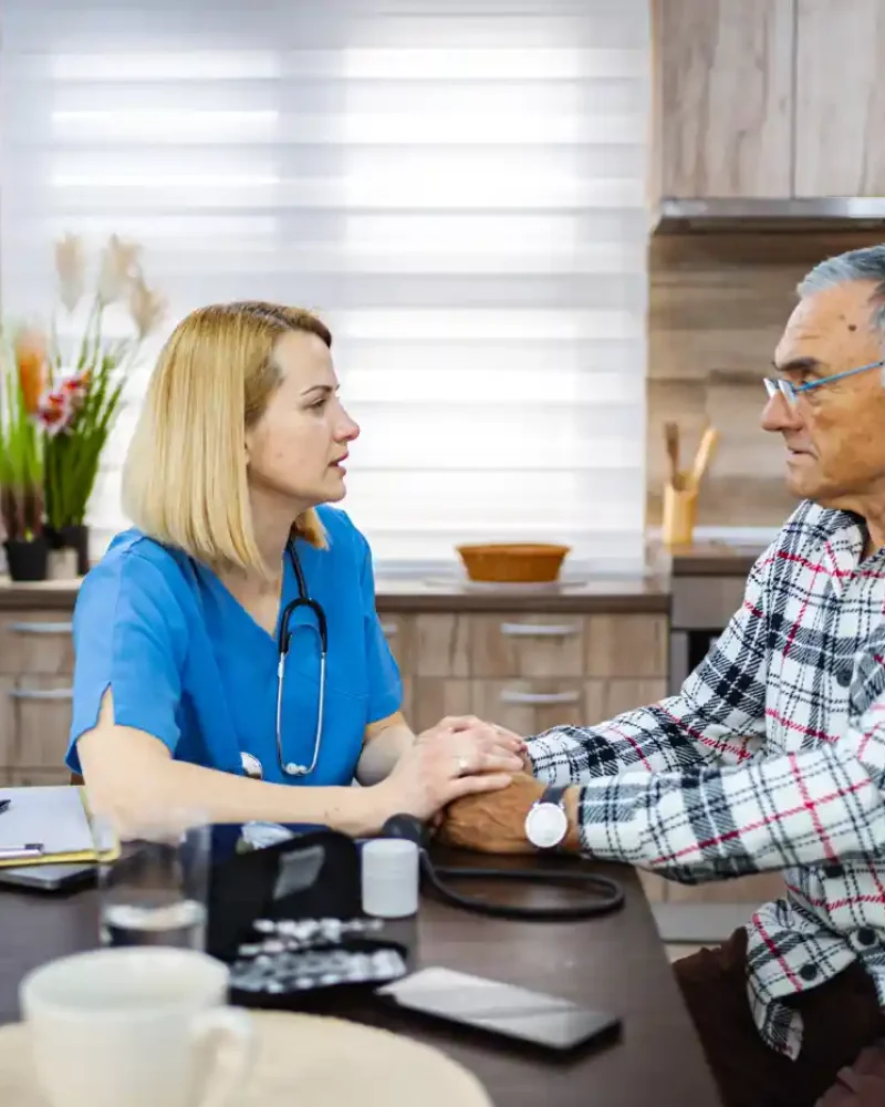 A female healthcare worker holds hands with an older man during a consultation at a kitchen table, with medical equipment and documents nearby.