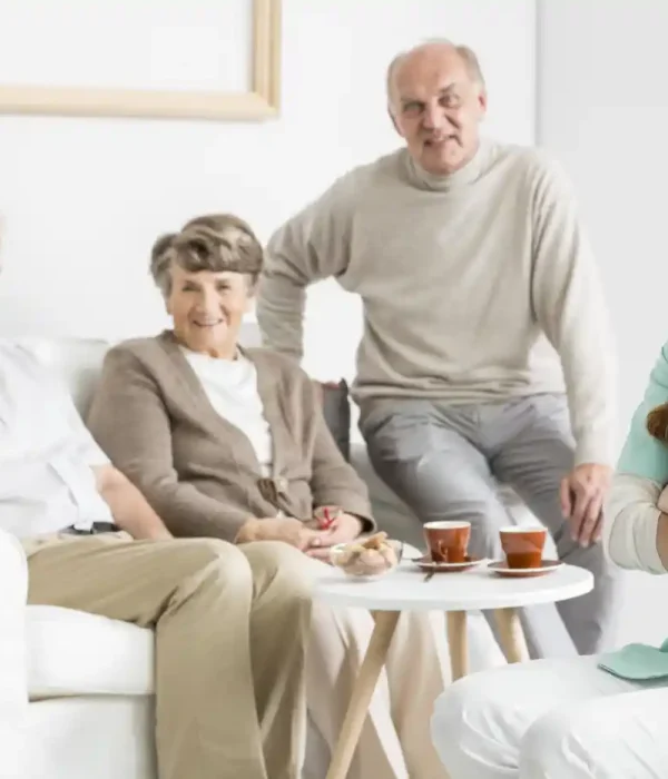Three elderly people sit together on a couch and chair in a living room while a young female caregiver from home care services stands in front, smiling, with tea cups on the table beside them.