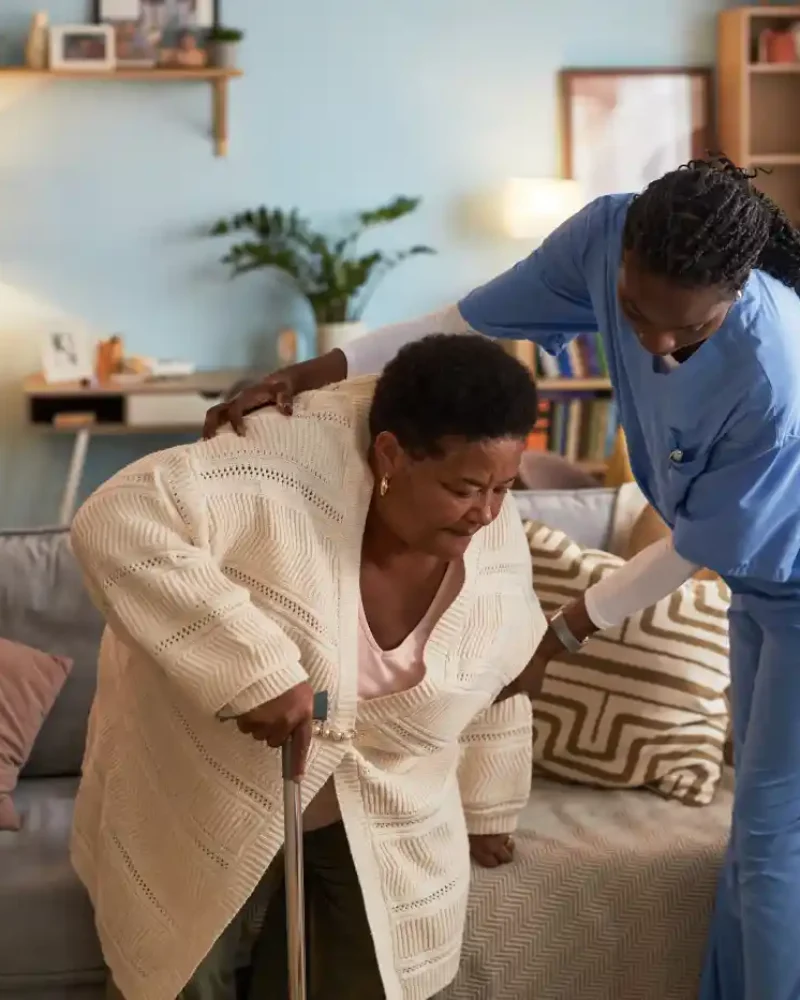 A healthcare worker provides in home care in Columbus, Ohio, assisting an older woman with a cane as she stands up from a couch in a well-lit living room.