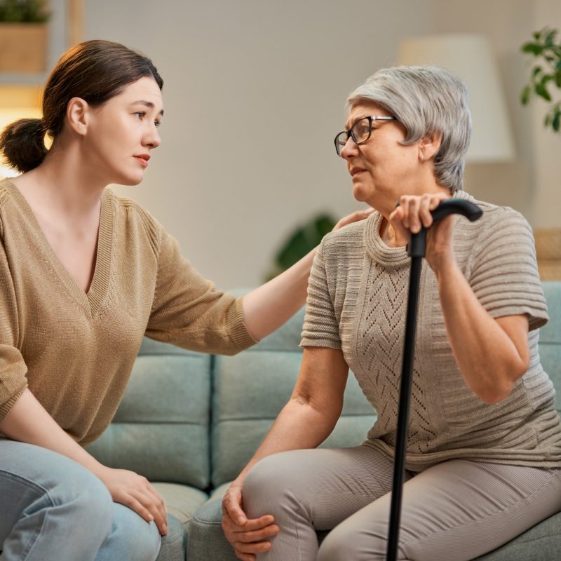 A woman is sitting on a couch talking to an older woman with a cane.