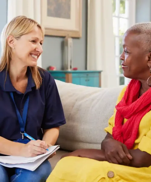 A woman in a navy shirt holding a clipboard smiles while discussing programs with another woman in a yellow outfit and red scarf, as they sit together on a sofa in a bright room.