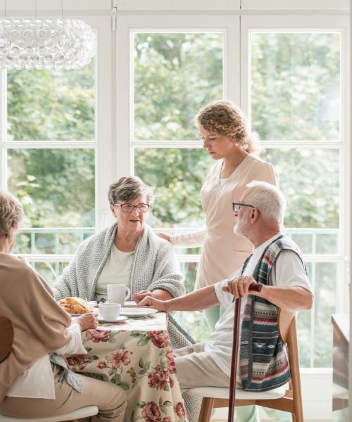 Group of senior friends with helpful carer sitting together at the table at nursing home dining room