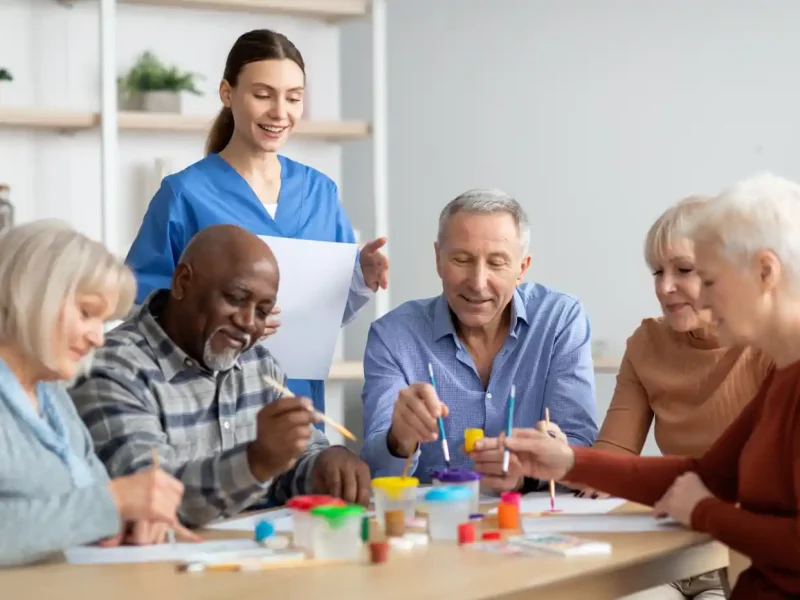 A group of older adults paints together at a table, assisted by a smiling caregiver in blue scrubs standing nearby.