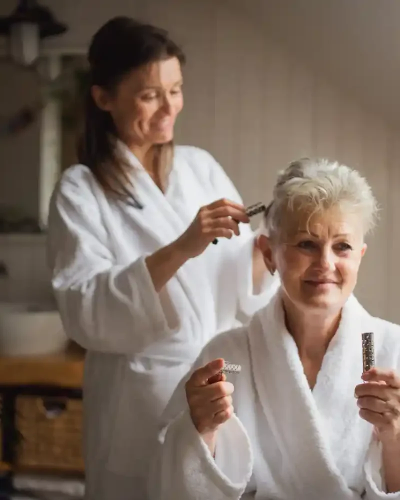 An older woman in a white robe sits and holds hair products while another woman in a robe styles her hair in a bright bathroom, highlighting the personal touch of in home care Columbus Ohio offers.