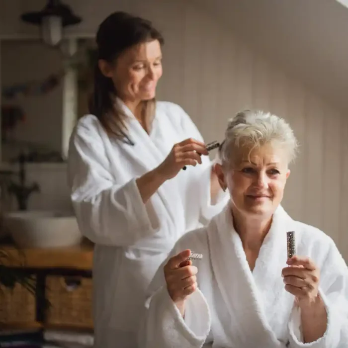 An older woman in a white robe sits and holds hair products while another woman in a robe styles her hair in a bright bathroom, highlighting the personal touch of in home care Columbus Ohio offers.