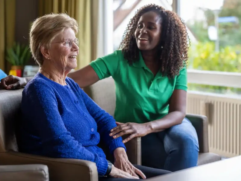 An older woman sits on a chair smiling while a younger woman in a green shirt, visiting for post hospital care, sits beside her talking and smiling in a bright living room.