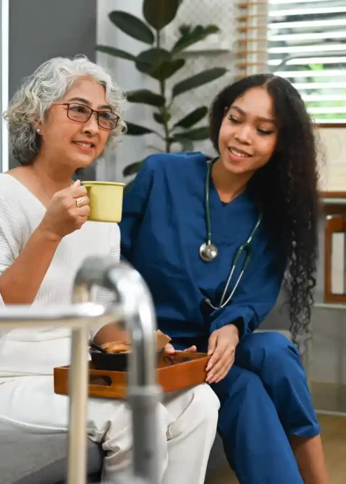An older woman sits on a couch holding a cup, while a nurse in blue scrubs—providing post hospital care—sits beside her, smiling. A walker and a tray with food are in the foreground.