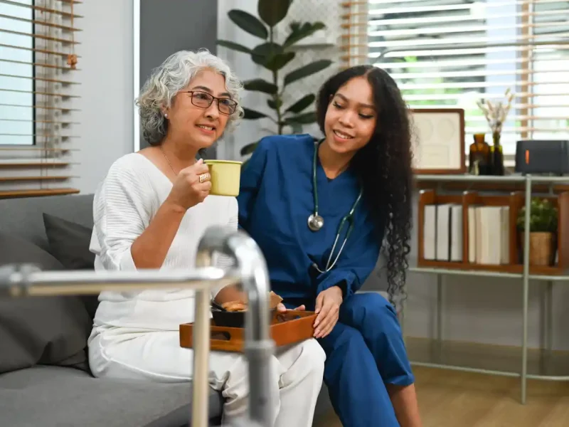 An older woman sits on a couch holding a cup, while a nurse in blue scrubs—providing post hospital care—sits beside her, smiling. A walker and a tray with food are in the foreground.