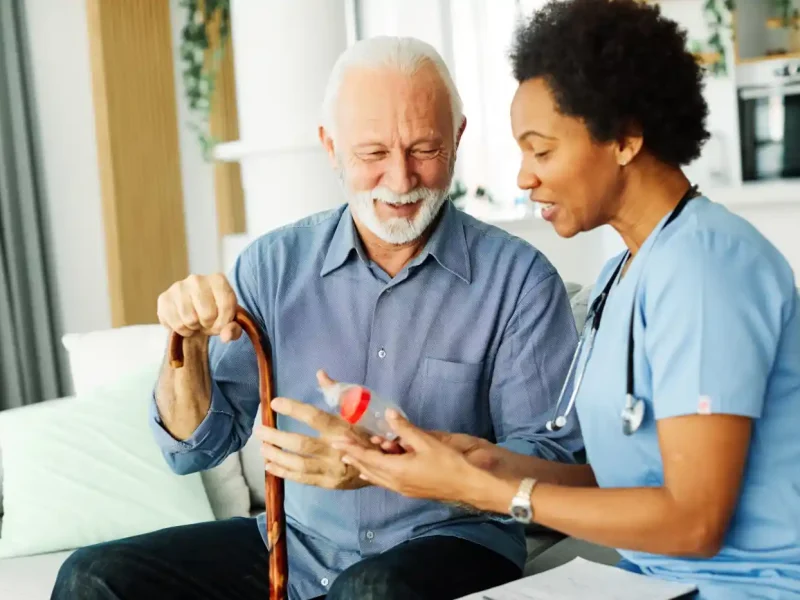A nurse in blue scrubs explains post hospital medication to an elderly man holding a cane, both seated indoors and smiling.