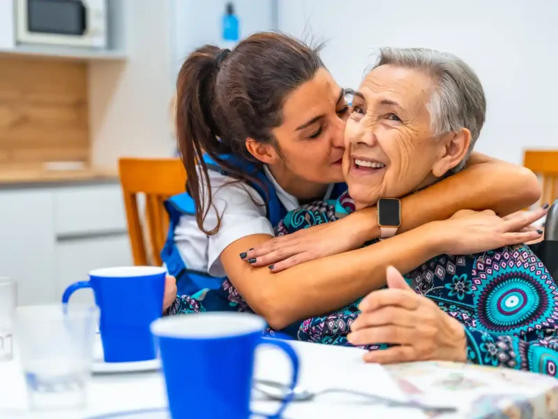 A young woman hugs and kisses an elderly woman, both smiling, while sitting at a table with blue cups in a bright kitchen—cherishing a joyful Post Hospital recovery moment together.