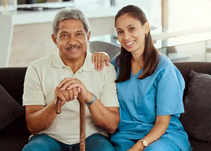 An older man with a cane sits on a sofa next to a smiling woman in blue scrubs, who has her hand on his shoulder, providing compassionate Alzheimer's and dementia care.