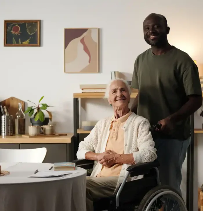 An elderly woman in a wheelchair sits at a table, smiling, while a man stands behind her in a home setting filled with plants, shelves, and wall art—highlighting the warmth of community programs that support seniors.