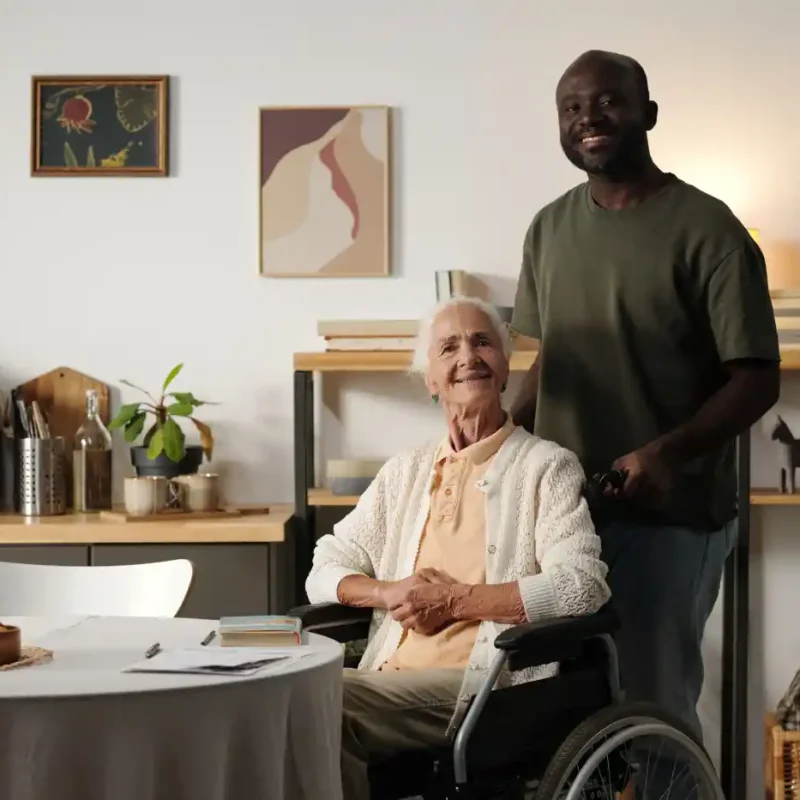 An elderly woman in a wheelchair sits at a table, smiling, while a man stands behind her in a home setting filled with plants, shelves, and wall art—highlighting the warmth of community programs that support seniors.