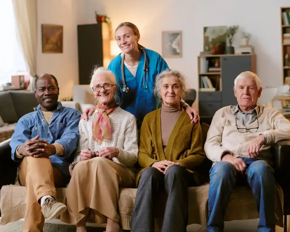 Four seniors sit on a couch with a healthcare worker standing behind them in a well-lit living room, all facing the camera—showcasing the warmth and trust that answer the question, "Why Us?.