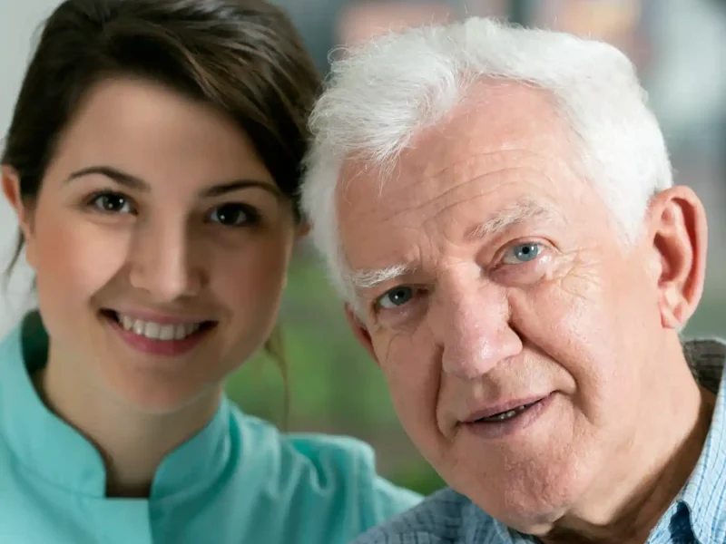 A young woman and an elderly man are posing closely together indoors, both smiling at the camera—a heartwarming image that captures the genuine connections behind Why Us.