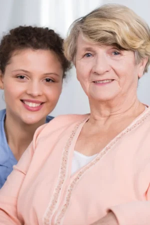 An elderly woman in a peach cardigan sits in front of a young woman with curly hair, both smiling at the camera, showcasing the warmth and companionship provided by Upper Arlington Home Care.