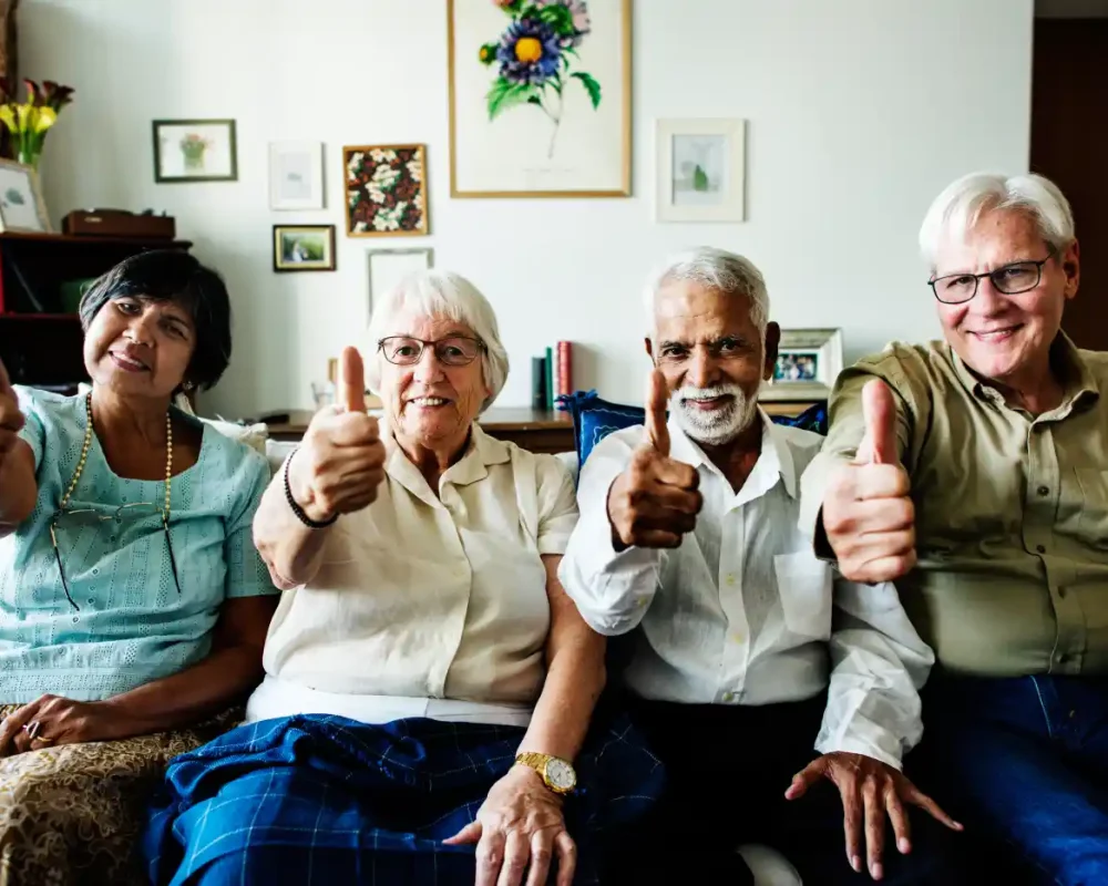 Four older adults sit on a couch indoors, smiling and giving thumbs-up gestures toward the camera, showing why us is the right choice for positivity and connection.