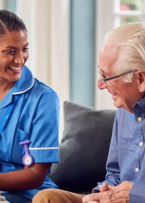 A nurse in blue uniform sits on a couch beside an elderly man, both smiling and conversing, with papers and a pen in front of them.