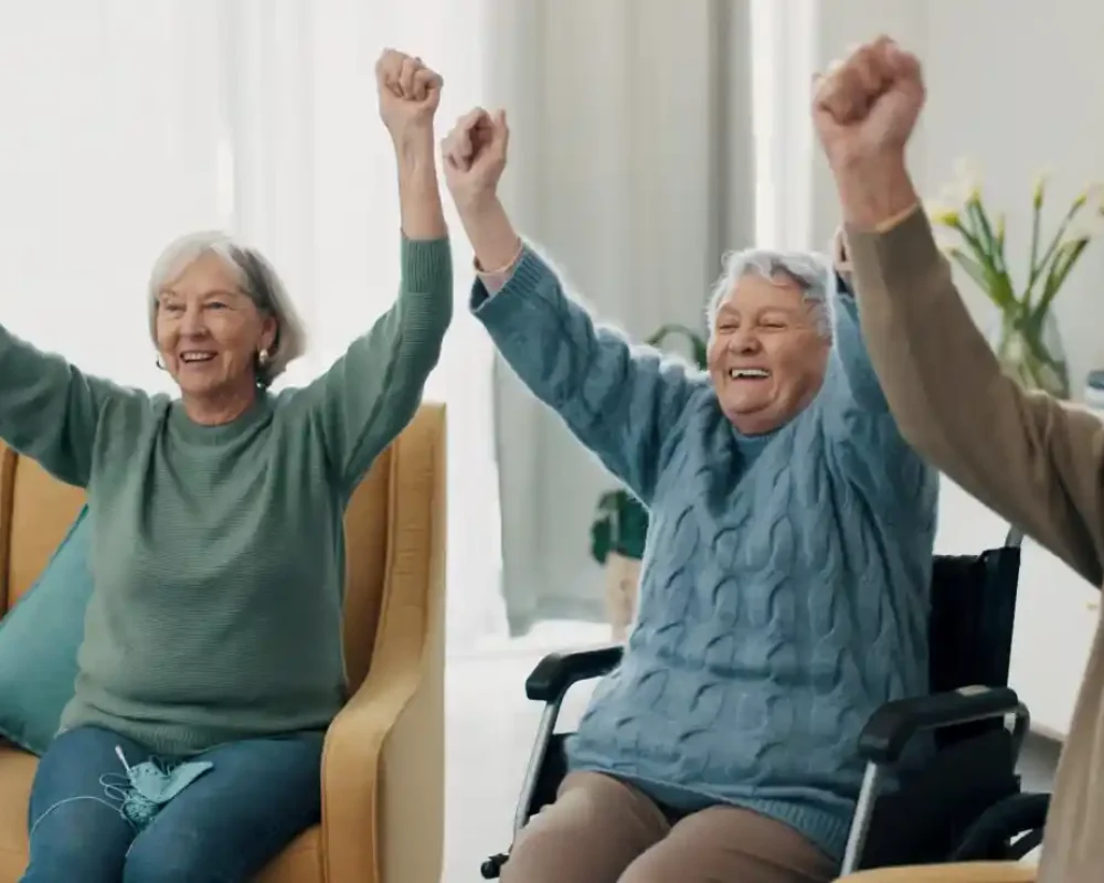 Three older adults sit side by side with arms raised and smiling, showing the enthusiasm and enjoyment that defines our Why Us spirit in a bright indoor setting.